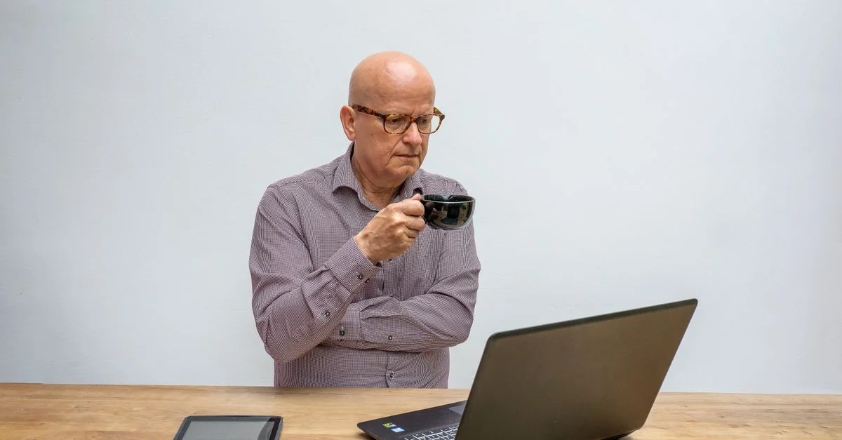 Un homme âgé pensif utilisant un ordinateur portable tout en sirotant un café, illustrant le travail à distance.
