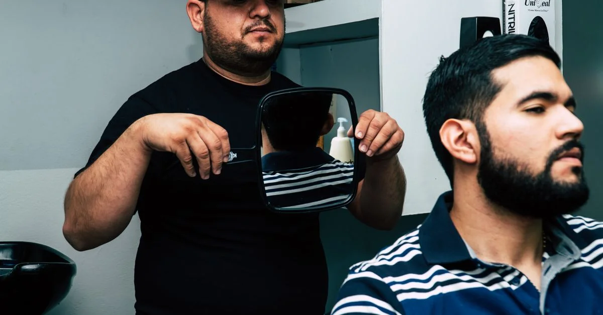 Un coiffeur portant une casquette montre un miroir à un client avec une barbe stylée dans un salon de coiffure moderne.