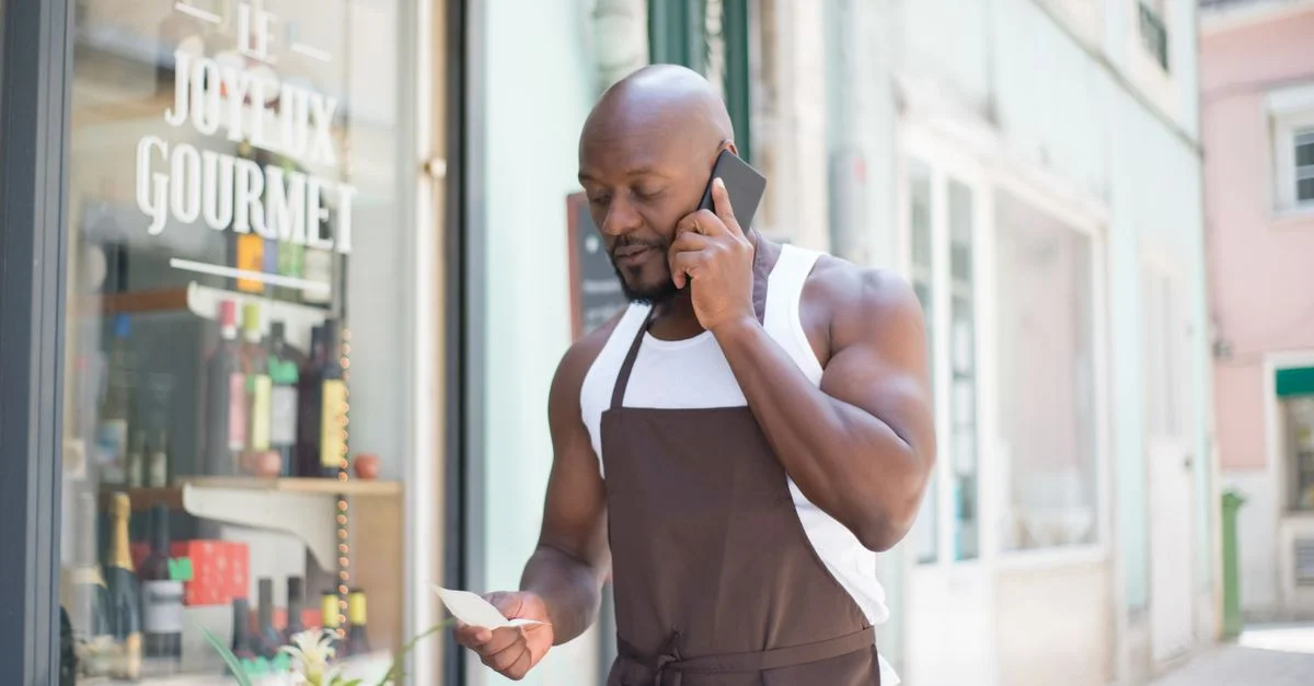 Homme en tablier passant un appel professionnel devant un magasin gourmet au Portugal, consultant des notes.