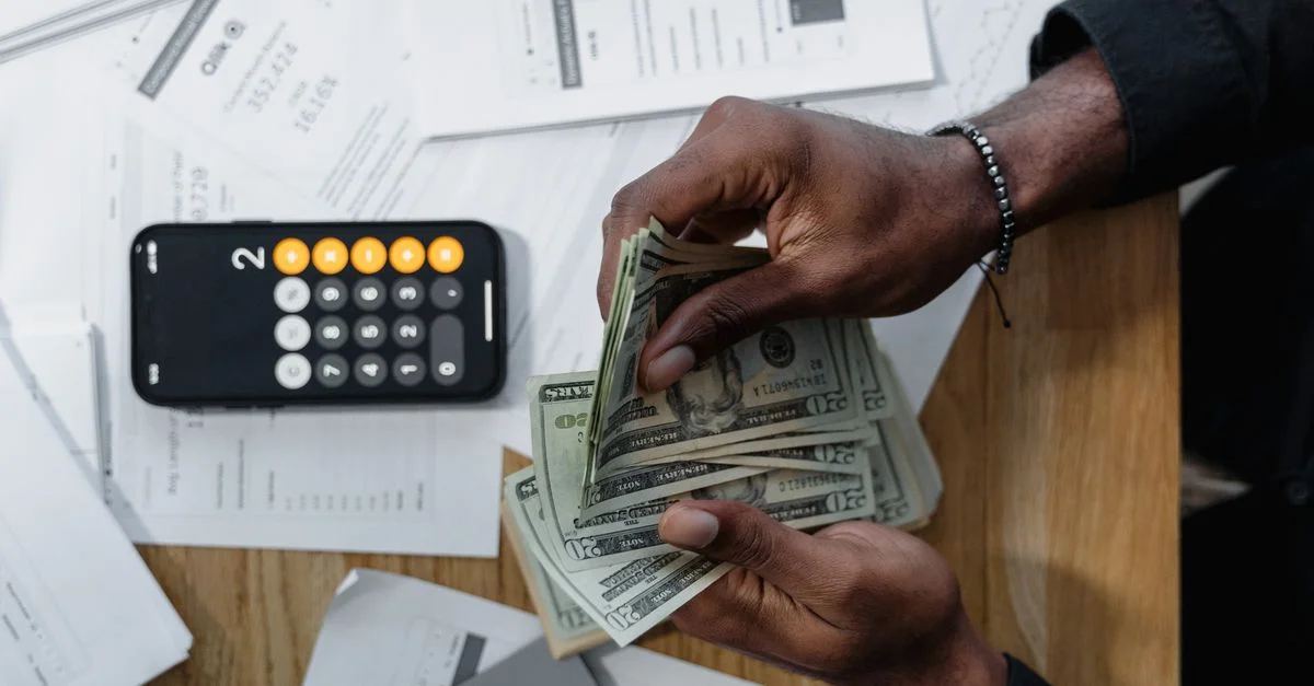 Personne comptant des billets de dollar sur des documents avec une calculatrice de smartphone sur le bureau.
